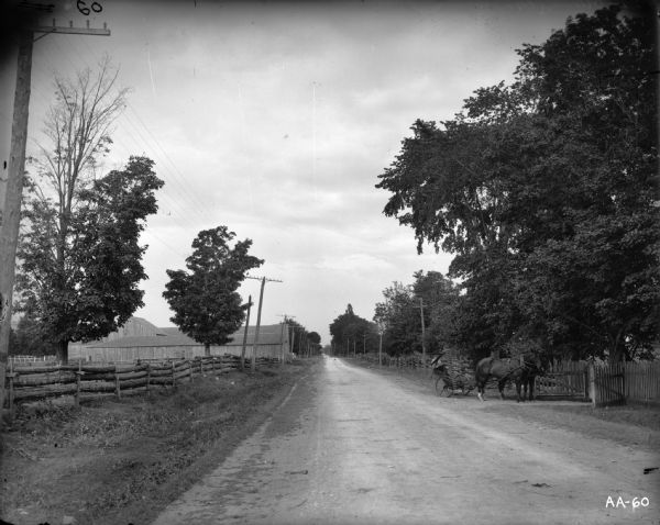 A farmer operating a horse-drawn mower entering a gate in a split-rail fence. A dirt road is leading off into the distance. There are also several wooden farm buildings along the side of the road.