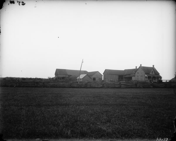 Scenic view across field of farmstead, including farmhouse, two large farm buildings, one small shed, and a split-rail fence. A family is posing on the porch and cows are grazing in a fenced-in area.