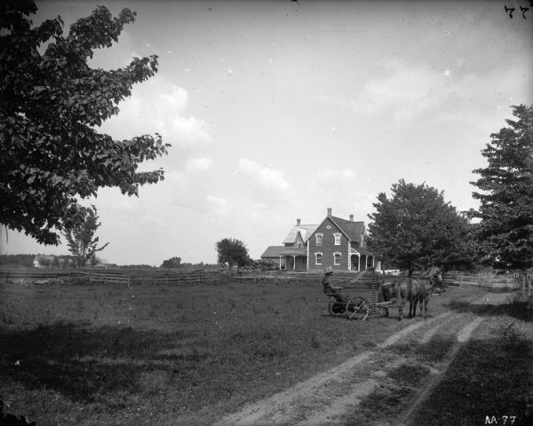 A farmer operating a horse-drawn mower enters a path leading to a brick farmhouse. The image contains architectural details, including decorative brickwork on the side of and around the windows of the farmhouse and gingerbread scrollwork (corbels) on the porch roof. There is also a split-rail fence in the image, and what appears to be a pile of debris next to the house.