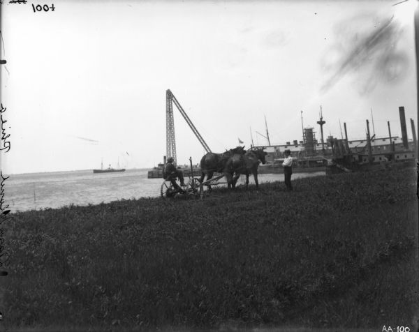 A man in work clothes is operating a horse-drawn mower in a patch of grass along a waterfront, while another man wearing a hat and tie is looking on. Ships, a dock, and a crane are on the shoreline. The words "League Island Phila" have been etched in the emulsion of the photograph along the left vertical edge. League Island was a naval shipbuilding yard on the Delaware River from the mid 19th to late 20th centuries.