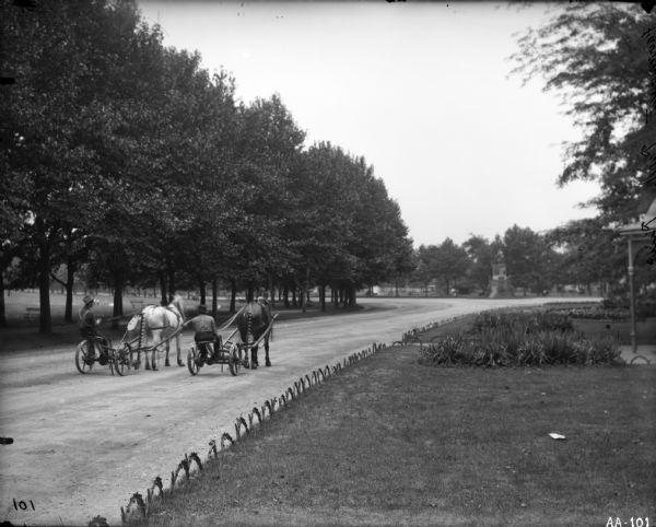 Two men are guiding horse-drawn mowers down a path in a park. There is a stand of tall trees along the left side of the path, and some low scalloped fencing along the right hand side. There is a statue, not easily visible, in the center of the path in the distance. The words "Fairmount Park Phila" are etched into the emulsion of the negative. Fairmount Park was the site of the 1876 Centennial Exposition.