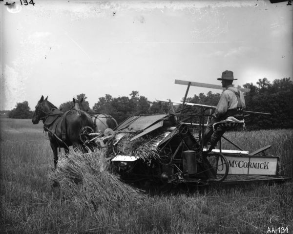 A farmer is operating a horse-drawn McCormick grain binder in a field. Many of the gear mechanisms in the rear of the binder are clearly visible in the image.