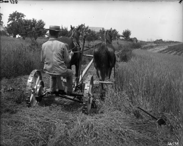 A farmer wearing a jacket and hat is operating a horse-drawn mower in a field. The horses are pulling the mower down a path that appears to have been mowed already; the grain on either side of the path is still unmowed. Farm buildings are in the distance.