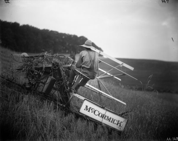 Rear view of a farmer operating a McCormick grain binder pulled by two horses on a hillside. In the background are hills and trees.