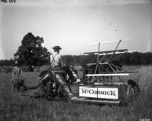 A farmer is operating a McCormick horse-drawn right hand open elevator grain binder (?) in a field while another man is gathering the bound sheaves of grain.