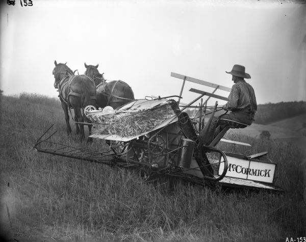 A farmer is operating what may be a McCormick open elevator (?) binder pulled by two horses in a field of grain on a gently sloping hillside.