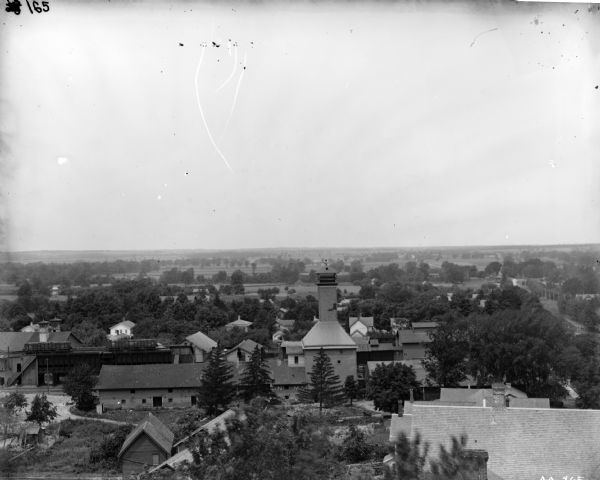 Elevated view of town and vicinity, perhaps taken from a hill, showing several buildings and the outlying rural area. Several of the buildings appear to be industrial rather than residential, as there are hopper cars on a railroad track running above a section of one building, and the letters "J.R.&" painted on the side of another.