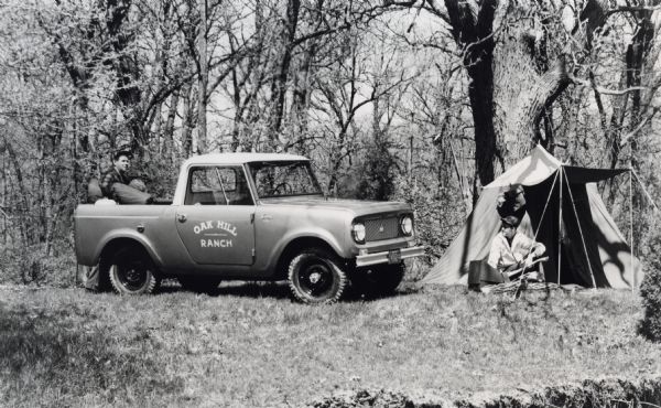 A man is unloading camping goods from the back of an International Harvester Scout parked in a wooded area, while another man is sitting in front of a tent nearby.