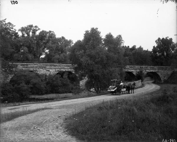 A man is driving a horse-drawn McCormick binder down a path that runs underneath a railroad bridge.