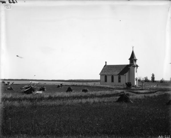 Field of grain and a McCormick horse-drawn binder in a field near a church. There are no horses attached to the binder. A person on a horse-drawn buggy is entering the yard of the church.