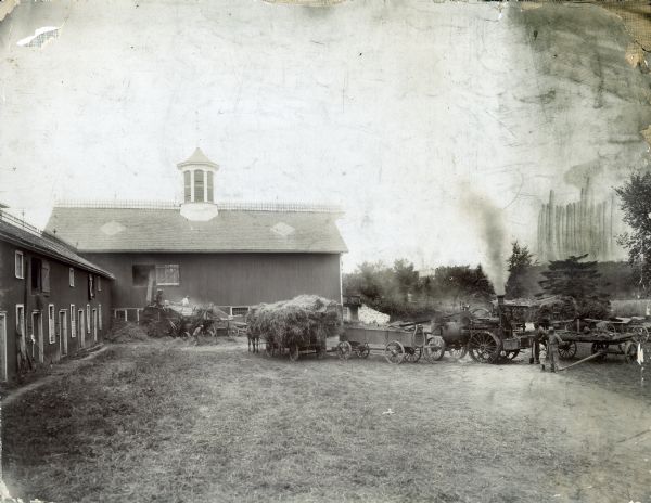 A group of men are working in front of a barn.  The equipment includes wagons, a steam engine, thresher, shovels, and extra wheels on the right.  One of the wagons has a large pile of hay loaded on it, and a horse's legs are behind the wagon on its left.  Three men are working with the machinery on the left. Two men and a young child are watching from the right.