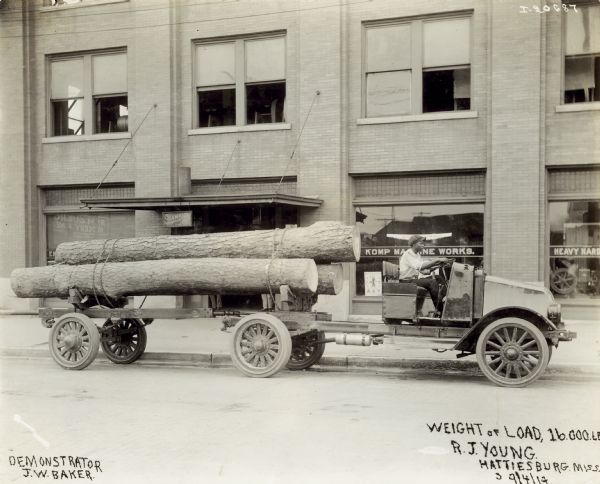 A man in a hat drives an International model "G" truck carrying logs with a wagon past a building. On the image is written: "Weight of load, "16.000.LB", as well as: "Demonstrator J.W. Baker", and under the weight: "R.J. Young, Hattiesburg, Miss."