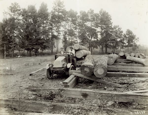 Two men sitting in an International model "G" truck pulling a wagon with logs tied together on top of it. Several logs are stacked on top of wood beams to the right. In the background is a line of trees. The man driving is African American. Both men are wearing hats.