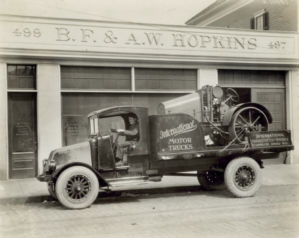 Man driving an International model "G" truck with an International 8-16 tractor tied to the bed. Behind the truck is a building for B.F. & A.W. Hopkins, Truck Dealers. The road is paved with bricks. The truck has no doors on the cab.