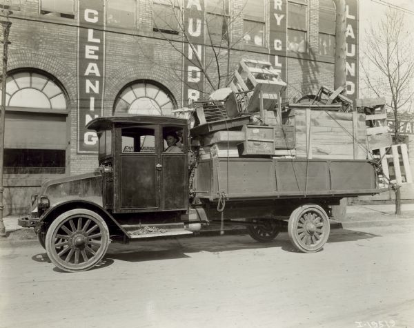A man sits in an International model "G" truck that has wood furniture and crates filling the bed. The building behind has signs advertising for a laundry.