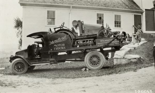 A man sits in an International model "G" truck carrying an International 8-16 tractor and other farm equipment, which is parked on a grassy hill next to a building. Rocks are piled in front of one of the rear wheels. A young girl stands on a stone retaining wall behind the truck.