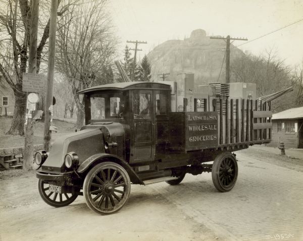 An International model "G" truck is in the process of turning from a brick-paved road onto a dirt drive. On the truck is written Latsch & Son Wholesale Groceries. In the bed of the truck are boxes of various items. Hanging from a post on the left is a sign for Wilkie's Garage and Machine Works. In the background is a very tall and rocky hill with some sparse bare trees.