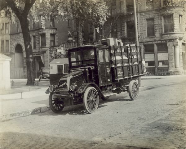 An International model "G" truck is near a curb on a brick-paved street, with many buildings in the background. The street corner is just behind the truck on the right. On the truck is written: "Peet stock Remedy Co." and "Peet Protection(?) Powder." In the bed of the truck are a variety of items.