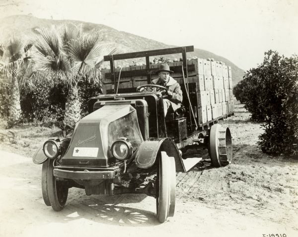 A man is driving an International model "G" truck which is turning on a road from a dirt path from what appears to be an orchard. Two palm trees are on the left. On the bed of the truck are crates of produce. The back wheel has a round and ribbed attachment.