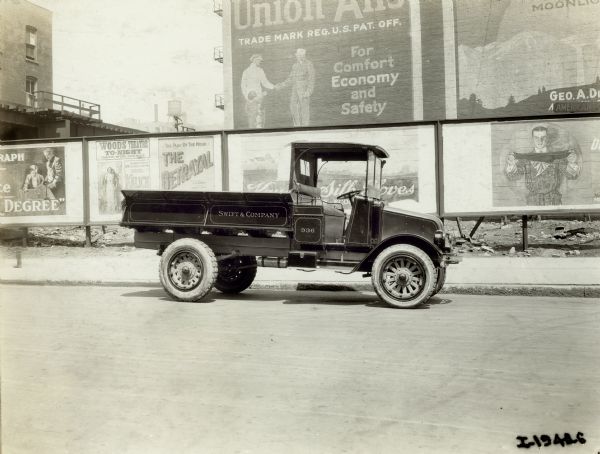 International model "G" truck with an empty bed sits parked along a curb.  On the side is written: "Swift & Company." Behind the truck is a line of billboards and buildings with advertisements for products and plays.
