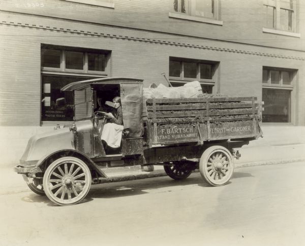 Two women sitting in the cab of an International model "G" truck parked next to a curb. The bed of the truck holds baskets, and a shovel. Written on the side is: "F. Bartsch, Stand No. 181, N.S. Market; Florist and Gardner". The window in the building behind the truck has the words "International Motor Trucks" in the lower corner on the left.