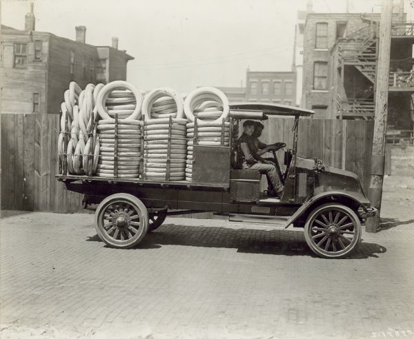 Two men are in sitting in the cab of an International model "G" truck, which is transporting a bed full of tires. They are on a brick-paved road. Behind them is a wood fence, and buildings in the background.