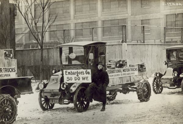 A man stands by an International model "G" truck, with one foot resting on the front wheel.  On the truck are two signs.  The one in front reads: "Embargoes Go, So Do We".  The other, on the side of the bed, reads "International Motor Trucks, From Akron to Philadelphia". The back tires of the have snow chains on them.