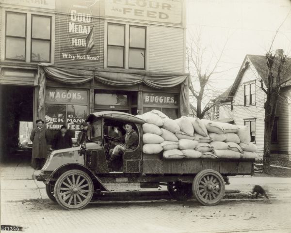 Two men are sitting in an International model "G" truck, parked next to a curb. Three men are standing against a storefront on the left. At the back of the truck is a dog on the street near the curb. On the bed is a "load 5 tons flour hauled from Chicago" (written below the image). In the background is a store selling flour, feed, wagons, and buggies. The vehicle is labeled as "F.C. Siekmann Truck" which is also the name on the shop window. To the right in the background is a house and some trees.