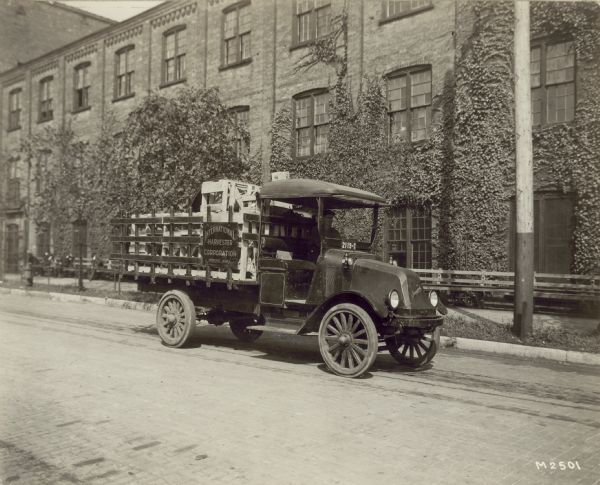 Original caption reads: "Model G, two-ton capacity, owned and operated by the International Harvester Corporation, Milwaukee, Wis, for cartage purposes, and which is giving very satisfactory service. Purchased during 1917." There is a man in the truck, which is parked along a street near a building, probably the Milwaukee Works (factory), covered in ivy. Wood crates are in the bed. In the background, men are resting on benches along the building. The sign on the side of the truck reads: "International Harvester Corporation Milwaukee Works."