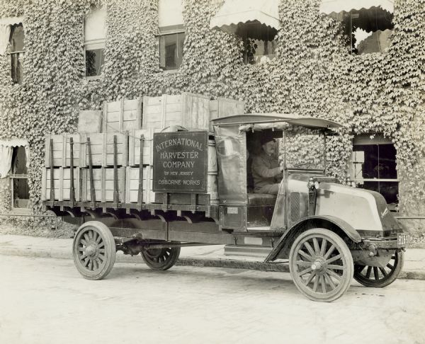 A man in an International model "G" truck with wooden boxes in the bed is parked outside of a vine-covered building. The building is most likely International Harvester's Osborne Works (the factory was later known as "Auburn Works"). A sign attached to the truck reads: "International Harvester Company of New Jersey Osborne Works".