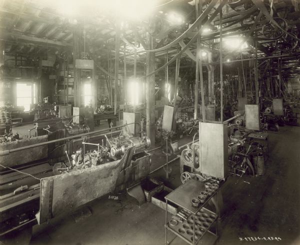 Slightly elevated view of machinery and machine parts in a large room. The machinery is belt driven from overhead. Light is flooding in from the windows and skylights in the ceiling. The factory manufactures 4 cylinder engines for general and farm work.