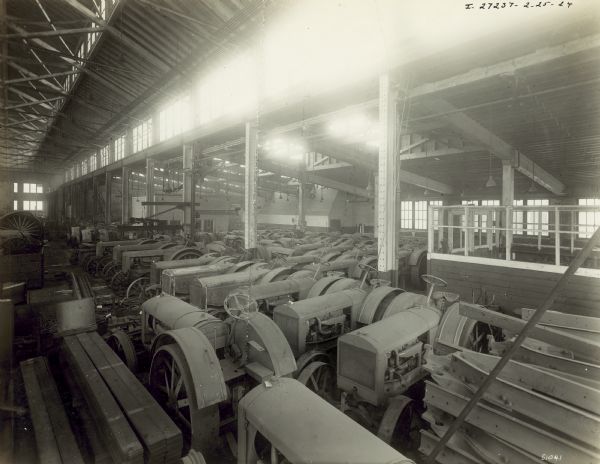 Stored in a large room are rows of tractors. Along the left and right corner are stacked beams. Light streams in from skylights in the ceiling. The factory manufactures 4 cylinder engines for general and farm work.