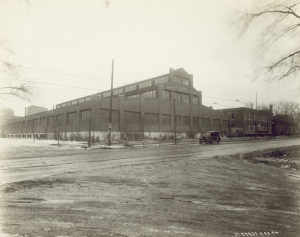 View across road towards a car driving down a cobblestone road in front of the factory. The factory manufactures 4 cylinder engines for general and farm work. Snow is on the ground. There are two other buildings to the right of the factory.