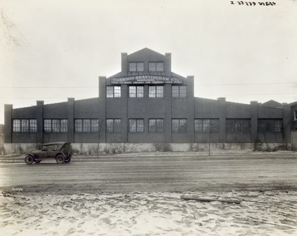 View across road towards a car parked on a brick-paved road outside the front of the factory. A sign on the front has the name of the factory and also reads: "Four Cylinder Engines For General Farm Work."