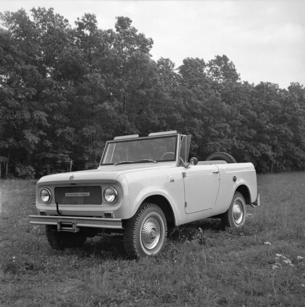 An International Scout truck, with top removed, parked in a field in front of a wooded area.