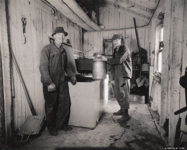 Two men are standing beside a McCormick-Deering Milk Cooler in a wood storage building. One man is wearing patched overalls and is holding a milk canister, while the other man is standing nearby.