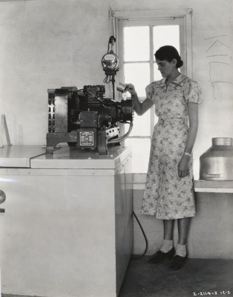 A woman holding a cup stands near a McCormick-Deering Milk Cooler near a window inside a building.