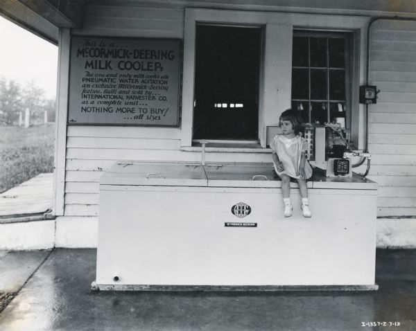 A two or three-year old girl (identified as Donnie May Childs) sits atop a McCormick-Deering 10-can milk cooler. The cooler sits under a roof outside a building. An advertisement hanging above the cooler reads: "This is a... McCORMICK-DEERING MILK COOLER The one and only milk cooler with PNEUMATIC WATER AGITATION an exclusive McCormick-Deering feature. Built and sold by... INTERNATIONAL HARVESTER CO. as a complete unit... NOTHING MORE TO BUY! ...all sizes". The photograph was taken at Telling Belle Vernon Farms in Novelty, Ohio.