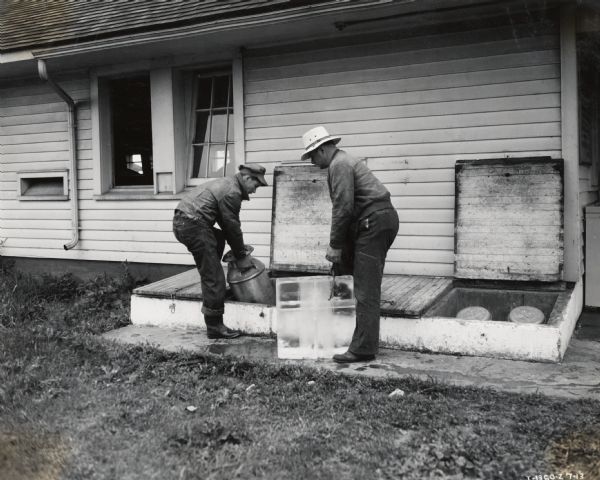 Two men stand on the side of a building.  On man is setting a milk canister into a bulkhead of a cellar, while the other stands to the side holding ice tongs to handle a large block of ice.  The photograph was taken at Telling Belle Vernon Farms.