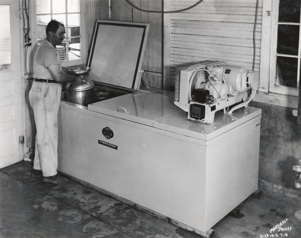 A man sets a milk canister into a McCormick-Deering 10-can milk cooler.  The photograph was taken at Holtex Farms in Dallas, Texas.
