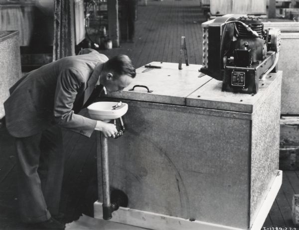 A man prepares to drink from a drinking fountain attached to a McCormick-Deering 4-can milk (water cooler), possibly at International Harvester's West Pullman Works. The cooler pictured was for display at a State Fair.