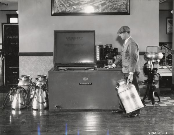 A man holds an empty milk container and looks at an open McCormick-Deering 6-can milk cooler on display in an International Harvester branch house or dealership. A photograph of McCormick Works hangs above the cooler.