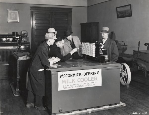 Five men are examining a 16-can McCormick-Deering Milk Cooler at an International Harvester dealership or branch house showroom. A man uses his pen to describe the features of the milk cooler's cooling mechanism.  The other four men (wearing hats) are looking at the cooler. A sign on the cooler reads "McCormick-Deering Milk Cooler 6-can capacity-cooling tank pneumatic agitator lubrication". A handwritten note also indicates the photograph came through the Jackson, Michigan Branch House.