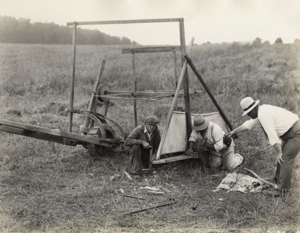 Men repair a reaper replica on the motion picture set of "Romance of the Reaper". The film was produced by International Harvester at Walnut Grove to celebrate the Reaper Centennial. Caption on back of photograph states: "1831 reaper. Replacing grain wheel with skid to correct historical error. Left to right: L.K. Swendsen, Deering Works expert; Dan Smith, expert from McCormick Works, and H.A. Kellar."