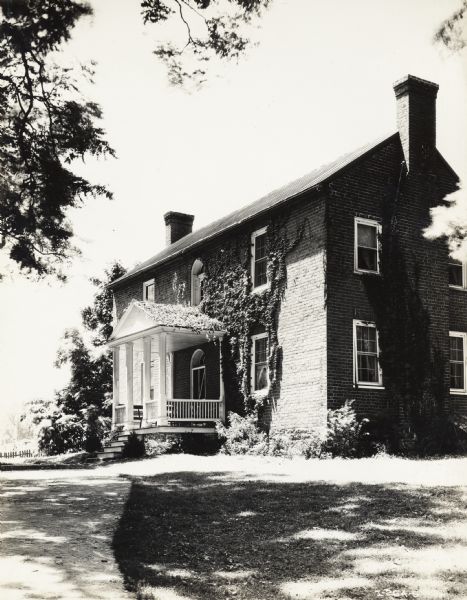 Production still for the Fox Hearst film "Romance of the Reaper". The film was produced by International Harvester at Walnut Grove to celebrate the Reaper Centennial. Caption on back of photograph states: "McCormick home prepared for filming. Note down-spouts removed, roof over porch covered with ivy, and large windowpanes which would show in picture, taped to resemble small panes."