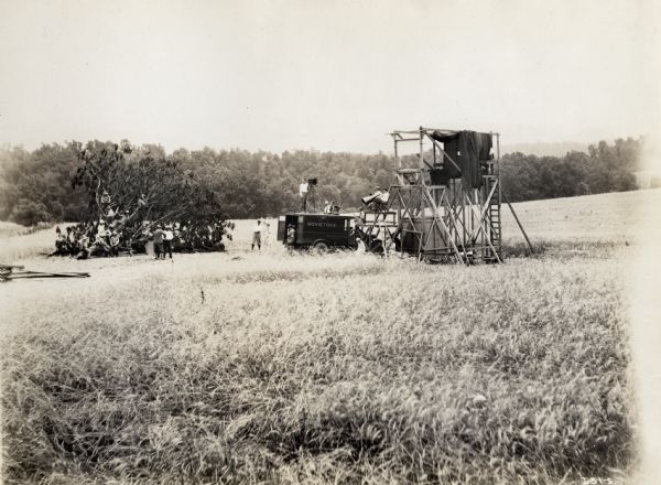 Production still for the Fox Hearst film "Romance of the Reaper". The film was produced by International Harvester at Walnut Grove to celebrate the Reaper Centennial. Caption on back of photograph reads: "Directing rehearsals for scene shot through glass. Group in fallen tree are local farm hands off location. Others are: Sanford B. White, Mrs. F.M. Somerville, Jr., E.F. McGovern standing on Movietone truck; H.A. Kellar and Mrs. Nathan seated on truck; Edgar L. Kaw and R.A. Chapman seated on platform."
