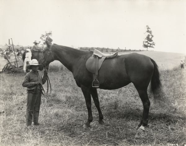 A photograph from the set of the Fox Hearst film "Romance of the Reaper". The film was produced by International Harvester at Walnut Grove to celebrate the Reaper Centennial. A local African American boy, Harry Alexander, is holding the reins of Cyrus Hall McCormick's (Del McDermid) horse. An African American woman, Clara Wilson, is standing in the background by a reaper.