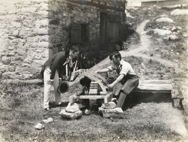A production still for the Fox Hearst film "Romance of the Reaper". The film was produced by International Harvester at Walnut Grove to celebrate the Reaper Centennial. Caption on back of photograph reads: "Cyrus Hall McCormick, (Del McDermid, professional actor) working on reaper outside forge shop, with William G. McCormick (Sam McClure, local boy) oldest of three younger brothers, assisting and looking on."