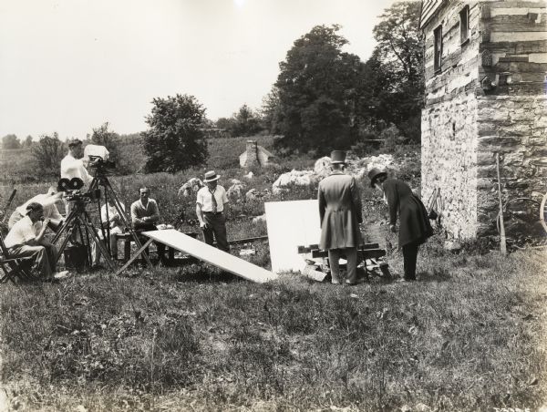 A photograph from the Fox Hearst film "Romance of the Reaper". The film was produced by International Harvester at Walnut Grove to celebrate the Reaper Centennial. Caption on the back of the photograph reads: "Fox Hearst crew directing action of Colonel James McDowell (J. Van Walton) and Captain William Massie (Vance McClure), who are discussing reaper. Reading left to right: R.A. Chapman, director; Herman Obrock, second cameraman; Charles Downs, head cameraman; Whitey, carpenter and one of their "grips"; "Sol," assistant cameraman, and Johnnie, who worked with the cameramen, usually handling the reflectors."