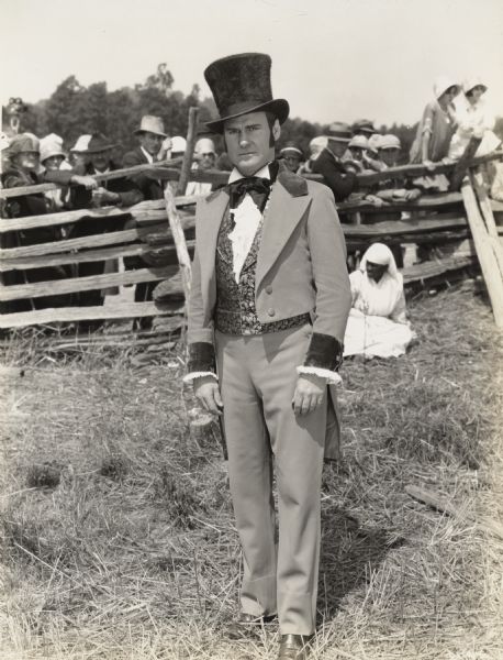 A production still from the Fox Hearst film "Romance of the Reaper". The film was produced by International Harvester at Walnut Grove to celebrate the Reaper Centennial. Del McDermid, the professional actor who played the role of Cyrus Hall McCormick, stands in the foreground wearing a suit and top hat. In the background, local African American woman Clara Wilson rests on the ground, and a group of individuals gather behind a fence.